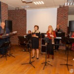 Na scenie biblioteki występują członkowie zespołu, akompaniujący śpiewaczce z tekstem piosenki wyświetlanym na ekranie za nimi.