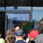 Grupa osób słucha prezentacji prowadzonej przed szklanym budynkiem podczas pikniku edukacyjnego.