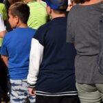 Grupa dzieci stoi tyłem, ubranych w kolorowe koszulki i czapkę.