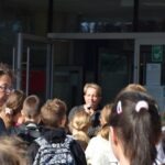 Grupa dzieci słucha prezentacji osoby dorosłej przed budynkiem.