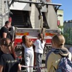 Grupa dzieci ogląda wnętrze strażackiego samochodu podczas pikniku edukacyjnego.