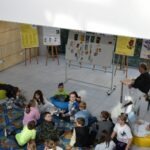 Grupa dzieci siedzi kręgiem podczas warsztatów edukacyjnych prowadzonych przez mężczyznę w pomieszczeniu z tablicami i plakatami.