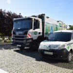 Na ulicy zaparkowany jest samochód dostawczy oraz śmietniarka oznakowane logo firmy zajmującej się gospodarką odpadami.