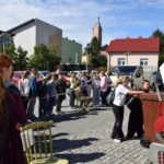 Grupa osób uczestniczy w pikniku edukacyjnym podczas Europejskiego Dnia Bez Samochodu na tle zabudowy miejskiej.