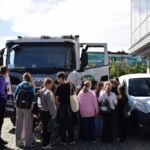 Grupa osób zgromadziła się przed ciężarówką do odbioru śmieci podczas wydarzenia promującego alternatywne formy transportu.