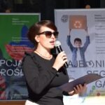 Kobieta przemawia do publiczności przez mikrofon, prezentując dokumenty na tle plakatów promujących ochronę środowiska.
