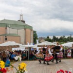 Zespół folklorystyczny prezentuje tańce regionalne podczas Święta Miodu w Płazie, z widoczną w tle nowoczesną architekturą kościoła.