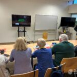 Osoby siedzące przy stole oglądają prezentację na ekranie telewizora w pomieszczeniu z tablicą.