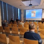 W sali konferencyjnej zgromadzeni uczestnicy słuchają prezentacji wyświetlanej na ekranie.