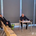 Dwóch mężczyzn siedzi w sali konferencyjnej, jeden przy stole z dokumentami, a drugi na krześle w tle.