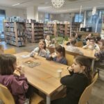 Dzieci siedzą przy dużym stole w bibliotece, otoczone regałami pełnymi książek.