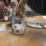 Dziecko maluje wzorem śnieżynki szklany lampion podczas warsztatów kreatywnych.