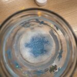 W szklanym pojemniku znajdują się niebieskie ozdobne elementy, prawdopodobnie związane z tematem podróży i przygód.