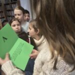 Dziecko trzyma otwartą książkę, a w tle widoczne są inne dzieci słuchające opowieści.