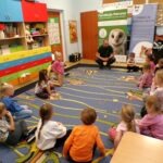 Grupa dzieci siedzi na dywanie podczas spotkania z przewodnikiem i prawdziwym jeżem Lucjanem.