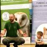 Mężczyzna w zielonej koszulce siedzi w pozycji lotosu przed banerem z wizerunkiem sowy, a obok niego stoi dziecko.