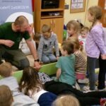 Mężczyzna w zielonej koszulce opowiada dzieciom o jeżu siedzącym na podwyższeniu.