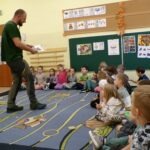 Mężczyzna czyta dzieciom książkę siedzącym na dywanie podczas warsztatów ekologicznych.