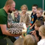 Mężczyzna z brodą prezentuje jeża Lucjana grupie zainteresowanych dzieci podczas warsztatów ekologicznych.