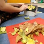 Dziecko przykleja fragmenty suchych liści do czerwonej kartki w trakcie tworzenia domku dla jeża.