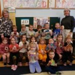 Grupa przedszkolaków stoi przed tablicą z literami, trzymając kolorowe domki dla jeży.