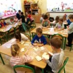Uczniowie przedszkola pracują przy stolikach, dekorując elementy domków z papieru i liści.