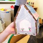 Ręka trzyma model domku z drewna, ozdobionego kolorowymi rysunkami kwiatów i roślin.