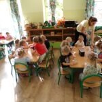 Dzieci siedzą przy stolikach podczas warsztatów plastycznych, tworząc figurki z drewna i liści.