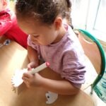 Dziecko maluje drewnianą figurkę jeża za pomocą kredki na białym papierze.