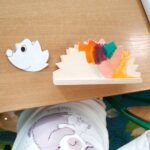 Na drewnianym stole leżą dwie figurki: biała ryba oraz jeżyk złożony z kolorowych elementów.