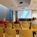 Uczestnicy warsztatów siedzą w sali, skierowani w stronę ekranu projekcyjnego z wyświetlaną prezentacją.