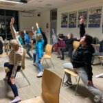 Uczestnicy zajęć wykonują ćwiczenia jogi siedząc na krzesłach w sali bibliotecznej.