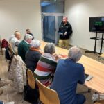 Grupa starszych osób siedzi przy stole podczas prezentacji, a mężczyzna w tle prowadzi wykład.