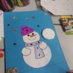 Dziecko stworzyło rysunek przedstawiający bałwanka z fioletową czapką i niebieską szalikiem na błękitnym papierze.