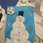 Dziecko stworzyło pracę plastyczną przedstawiającą bałwanka z koralikami i innymi ozdobami na niebieskim tle.