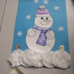 Dzieci stworzyły bałwanka z papieru i materiałów plastycznych na tle niebieskiego arkusza z płatkami śniegu.