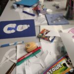 Na stole znajdują się kartki z rysunkami, kredki, mazaki oraz stempelki, co sugeruje organizację zajęć plastycznych dla dzieci.