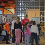 Grupa dzieci słucha opowieści prowadzącej zajęcia w bibliotece, otoczona elementami wystroju nawiązującymi do podróży.