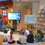 Dzieci siedzą na dywanie, skupione na projekcji wideo wyświetlanej na ekranie telewizora, a obok stoi tablica z mapą świata.