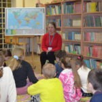 Nauczycielka w czerwonej bluzie opowiada dzieciom siedzącym na podłodze, prezentując mapę świata zawieszoną nad tablicą.