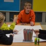 Dzieci siedzą przy stole, rysując kredkami na dużym arkuszu papieru w trakcie zajęć plastycznych.