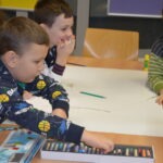 Dzieci siedzą przy stole, malując kredkami na dużym arkuszu papieru.