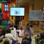 Grupa dzieci siedzi na dywanie, skupiona na projekcji wideo wyświetlanym na ekranie.