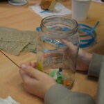 Dziecko wkłada kolorowe koraliki do szklanego słoika podczas warsztatów plastycznych.