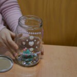 Dziecko trzyma ozdobiony cekinami i koralikami szklany słoik, który ma imitować lampion.