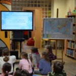 Grupa dzieci siedzi na podłodze, skupiona na prezentacji wyświetlanej na ekranie oraz mapie zawieszonej na fliparcie.