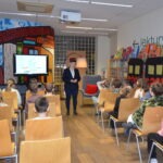 Prelegentka w marynarce przemawia do grupy dzieci siedzących na krzesłach w bibliotece, podczas gdy na ekranie wyświetlana jest prezentacja.