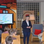 Mężczyzna w czapkach zorganizował prezentację dla dzieci siedzących przed ekranem, na którym widoczny jest schemat geograficzny.