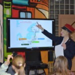 Nauczycielka w czapce prezentuje mapę na ekranie podczas zajęć dla dzieci.