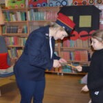 Kobieta w mundurze strażniczej wręcza dziecku ołówek podczas zajęć w bibliotece.